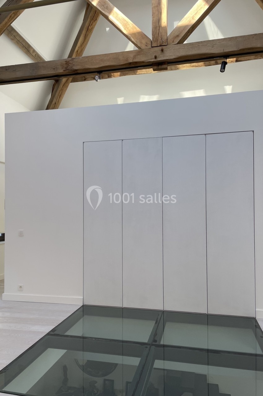 Plafond avec poutres apparentes en bois, mur blanc avec placards intégrés et sol en verre laissant voir un espace en dessous.
