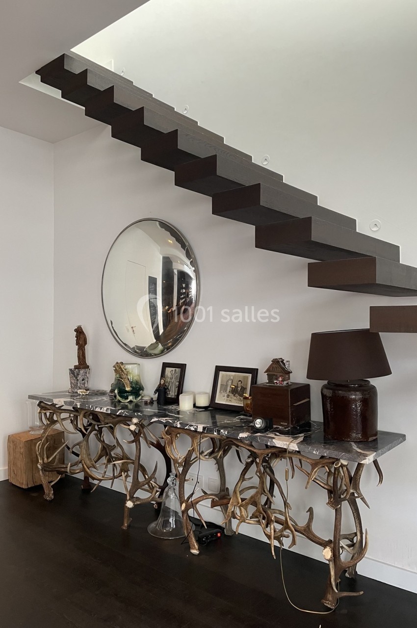 Escalier suspendu en bois sombre au-dessus d'une console décorée d'objets et d'un miroir rond, dans un intérieur moderne.