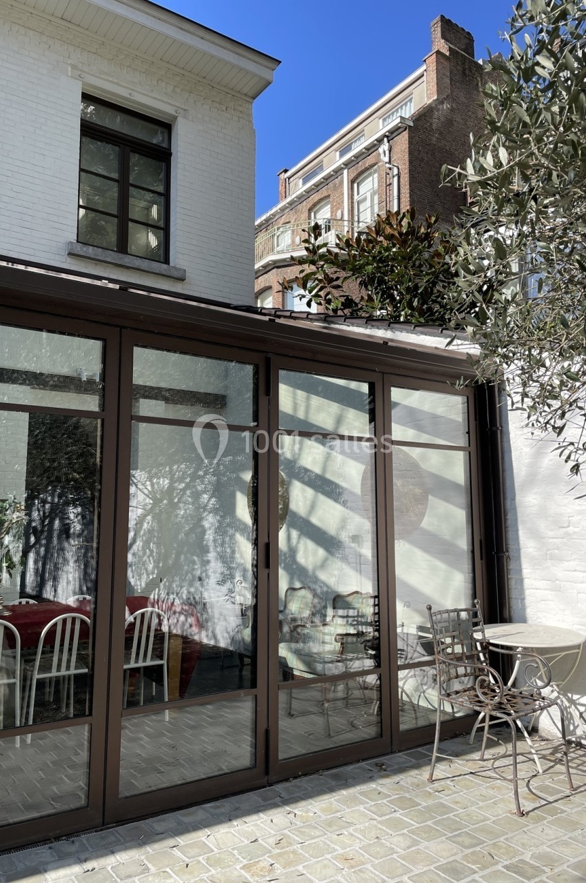 Verrière donnant sur une terrasse pavée avec table et chaises en métal, entourée de murs blancs et d'un arbre.