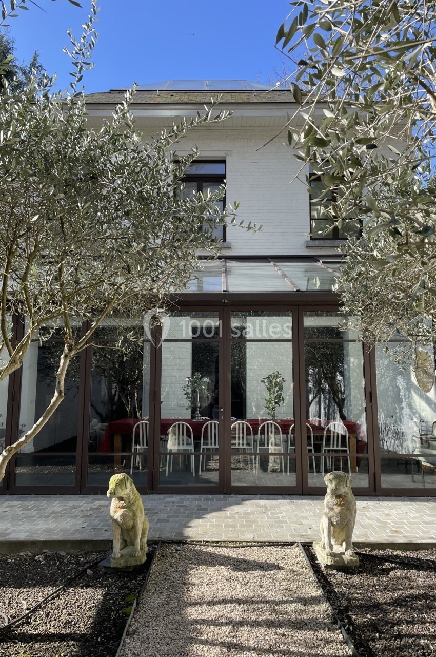 Façade d'une maison avec une véranda vitrée, entourée d'oliviers et ornée de deux statues de pierre.