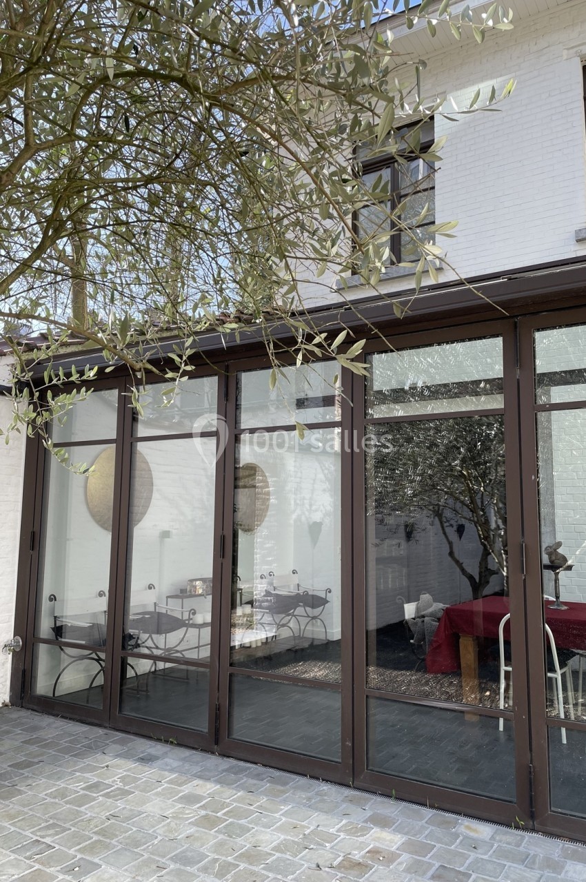 Baie vitrée donnant sur une salle à manger avec table rouge, vue depuis une terrasse pavée sous un arbre.