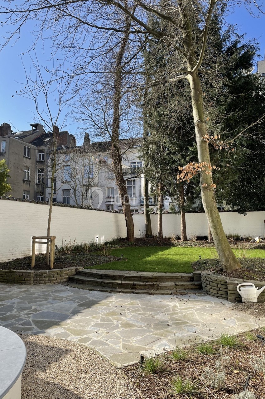 Jardin urbain avec pelouse, arbres, muret en pierre et sol pavé, entouré de murs blancs et d'immeubles en arrière-plan.