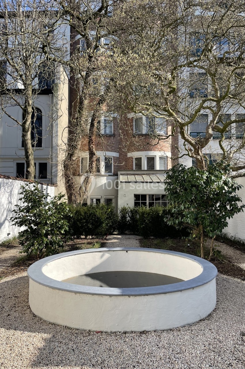 Cour intérieure avec un bassin circulaire blanc, entourée d'arbres, de buissons et de façades de bâtiments en arrière-plan.