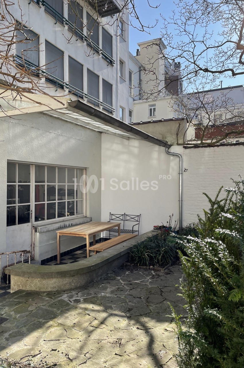 Cour intérieure avec table en bois, chaises métalliques, végétation et façade d'immeuble sous un ciel dégagé.