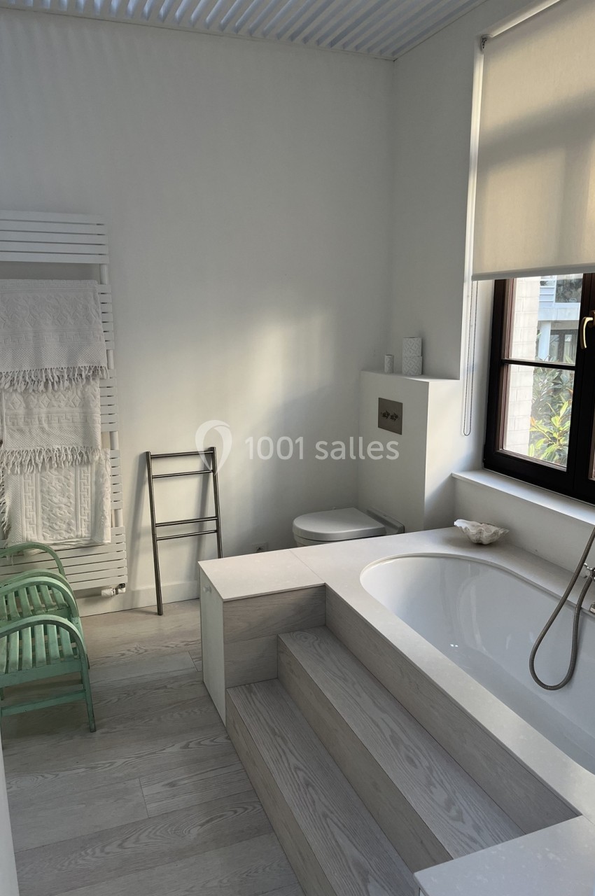 Salle de bain lumineuse avec baignoire encastrée, fenêtre, chaise verte, échelle et porte-serviettes.
