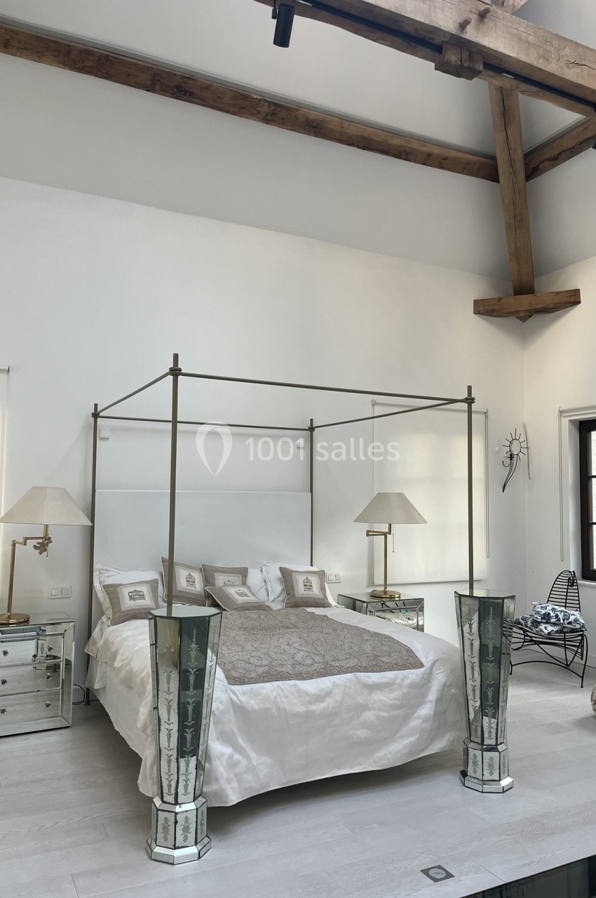 Chambre lumineuse avec lit à baldaquin, poutres apparentes, tables de chevet et grandes lampes décoratives.