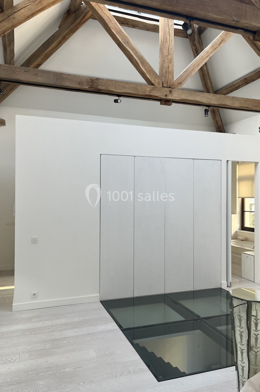 Plancher en verre transparent dans un intérieur moderne avec poutres apparentes en bois et murs blancs.