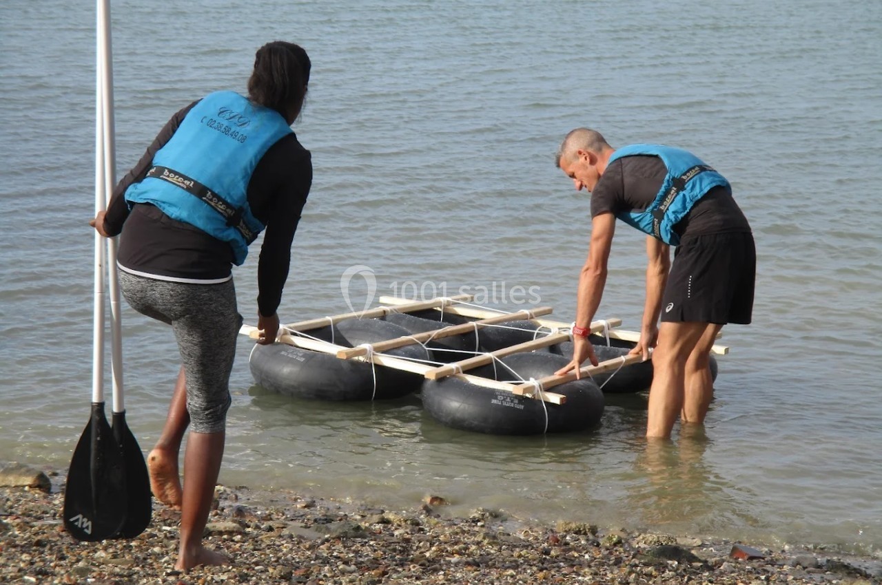 Deux personnes en gilets de sauvetage positionnent un radeau artisanal sur le rivage d'un plan d'eau calme.