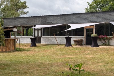 Miniature Location salle Saint-Guyomard (Morbihan) - Domaine de Ker Léon #4 Espace extérieur avec tables hautes noires sur une pelouse, devant un bâtiment vitré et un comptoir en bois.