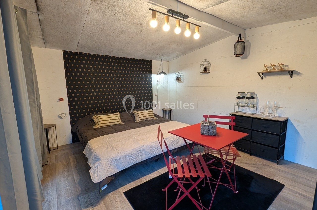 Chambre aménagée avec un lit double, une table rouge avec chaises pliantes, et une décoration murale sobre.