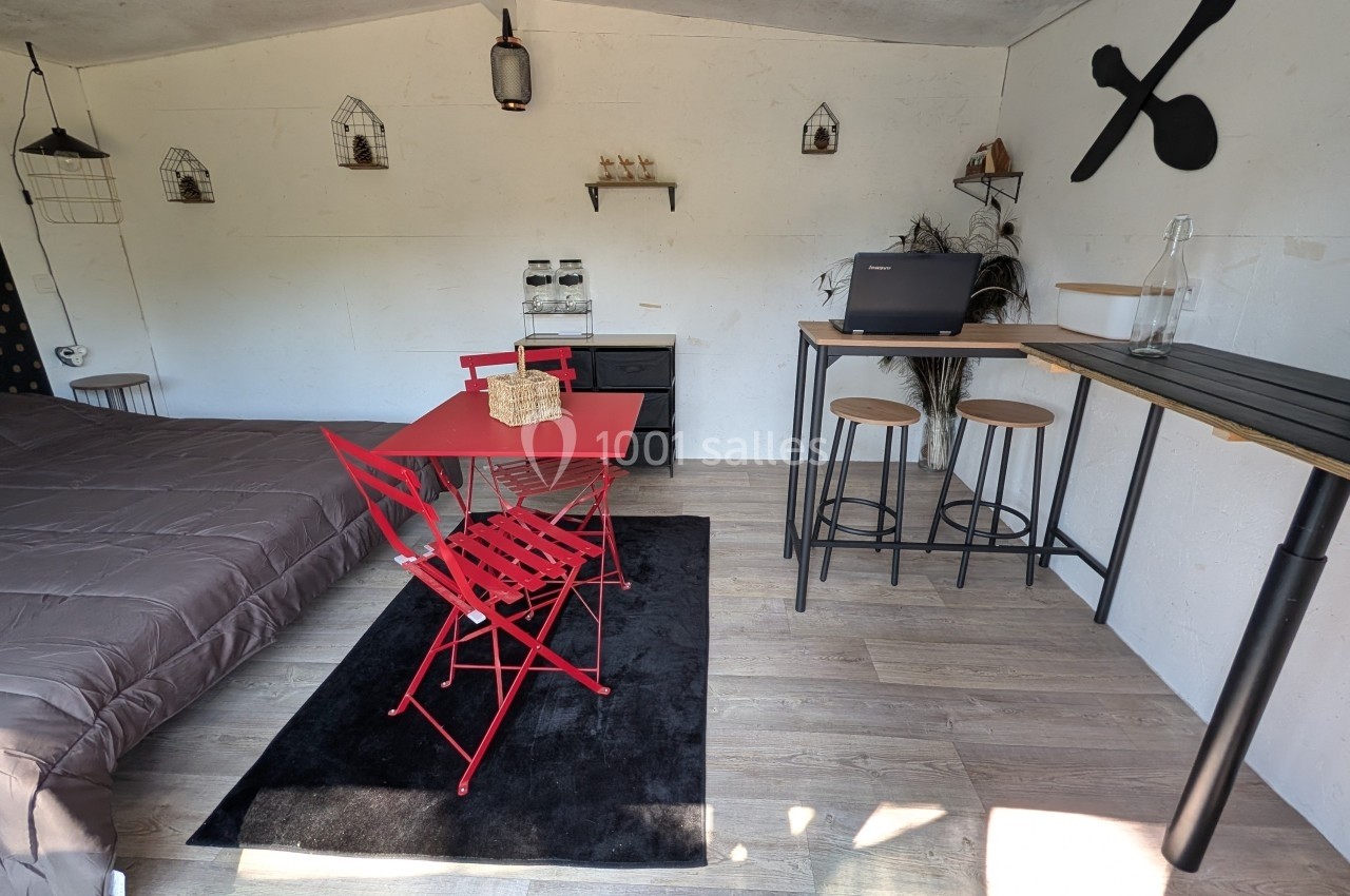Intérieur d'une petite pièce aménagée avec un lit, une table rouge, des tabourets hauts et une décoration minimaliste.