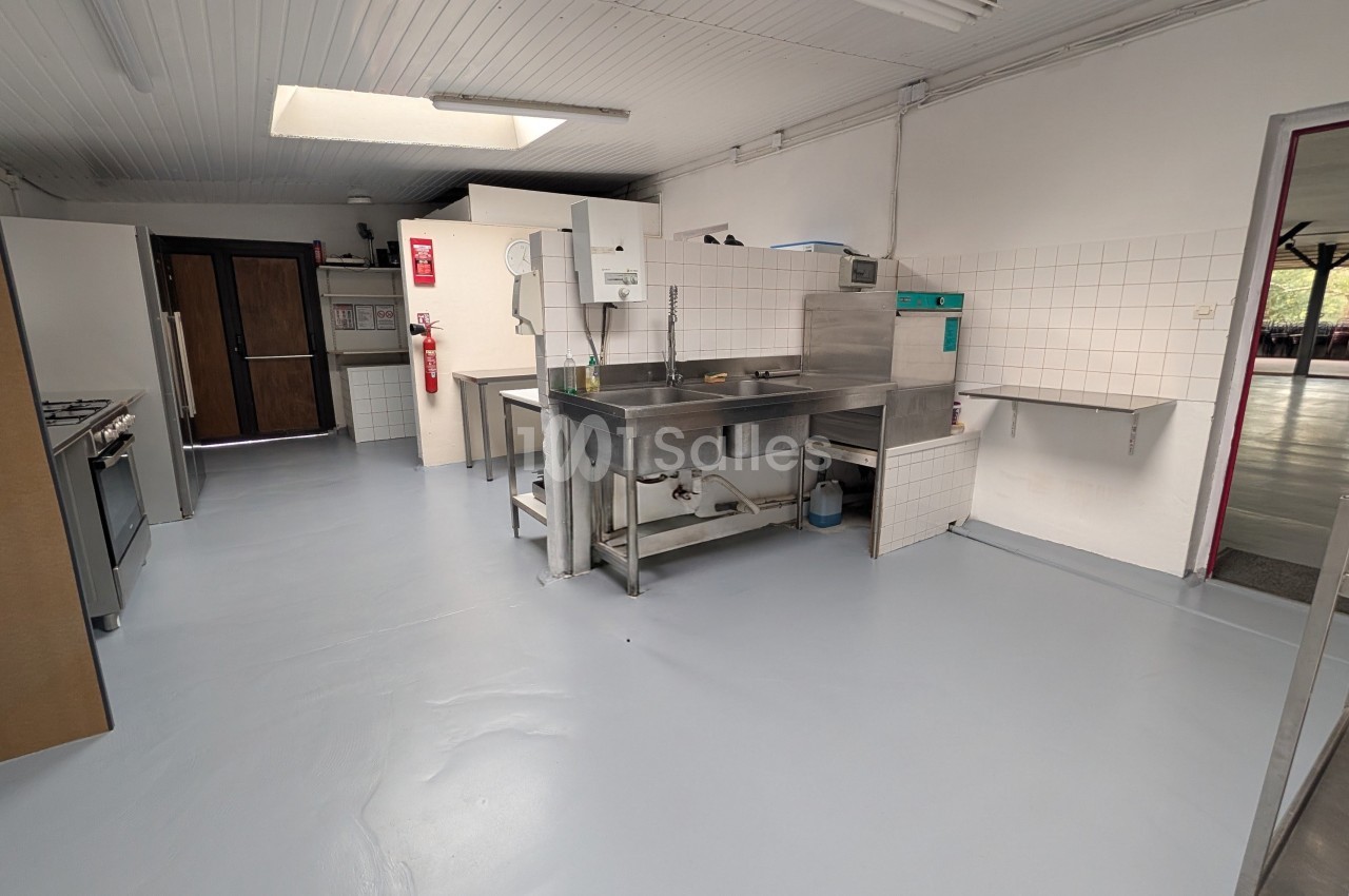 Cuisine professionnelle équipée d'un plan de travail central inox, réfrigérateur et rangements, sol gris clair.