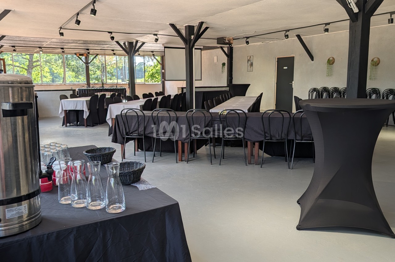 Salle intérieure avec tables dressées en noir et blanc, chaises noires, poutres apparentes, baies vitrées sur verdure.