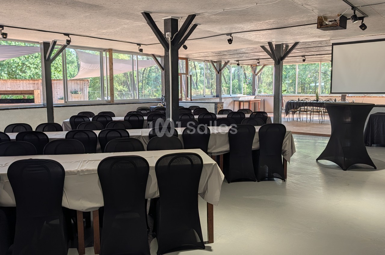 Salle intérieure spacieuse avec tables rectangulaires dressées, chaises noires, poutres apparentes et baies vitrées donnant sur espaces boisés.