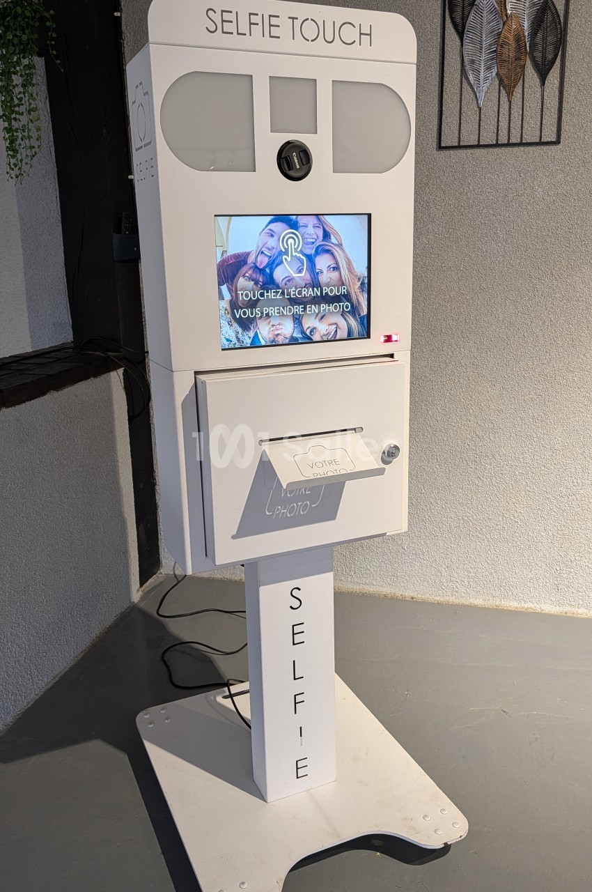 Photomaton blanc Selfie Touch avec écran tactile et bac de récupération des photos, installé en intérieur.