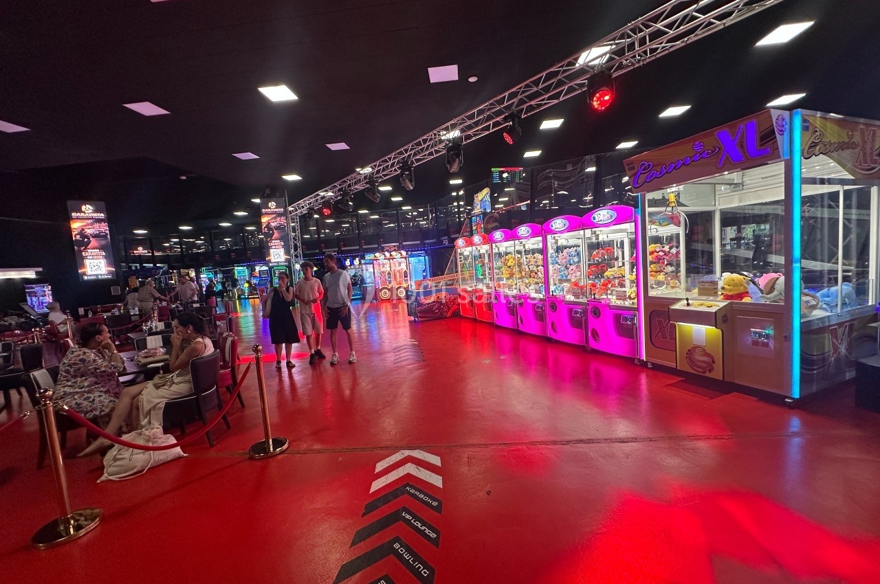 Salle de jeux avec machines à pinces lumineuses, sol rouge et visiteurs observant ou jouant.