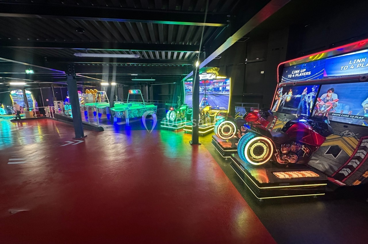 Salle d'arcade avec jeux vidéo lumineux, simulateurs de moto et tables interactives dans un espace sombre.