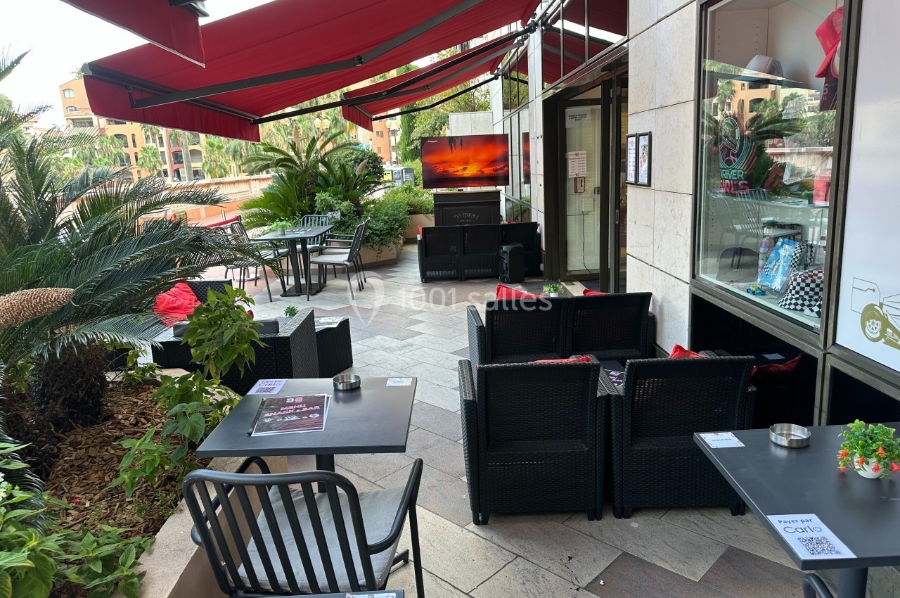 Terrasse extérieure d'un café avec tables, chaises et canapés sous des auvents rouges, entourée de plantes.