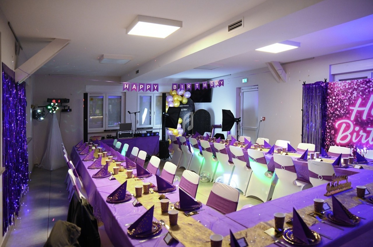 Salle décorée pour un anniversaire avec tables en U, nappes violettes, chapeaux pointus et guirlande ’Happy Birthday’.