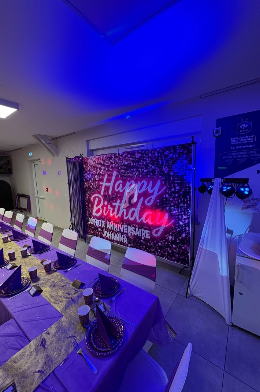 Salle décorée pour un anniversaire avec une longue table dressée en violet et une bannière lumineuse ’Happy Birthday’.