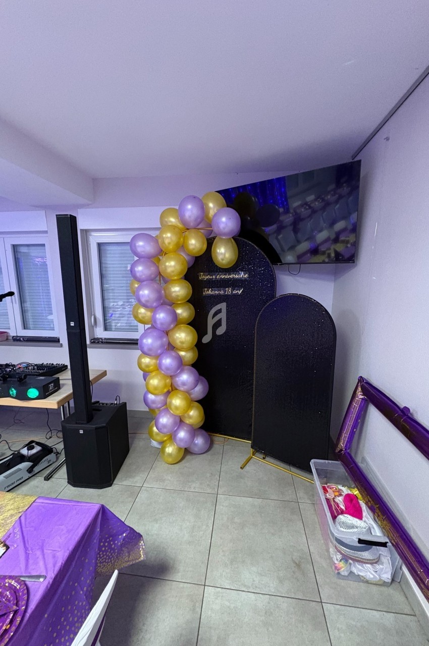 Guirlande de ballons violets et dorés décorant un coin de salle avec un écran et des équipements audio visibles.
