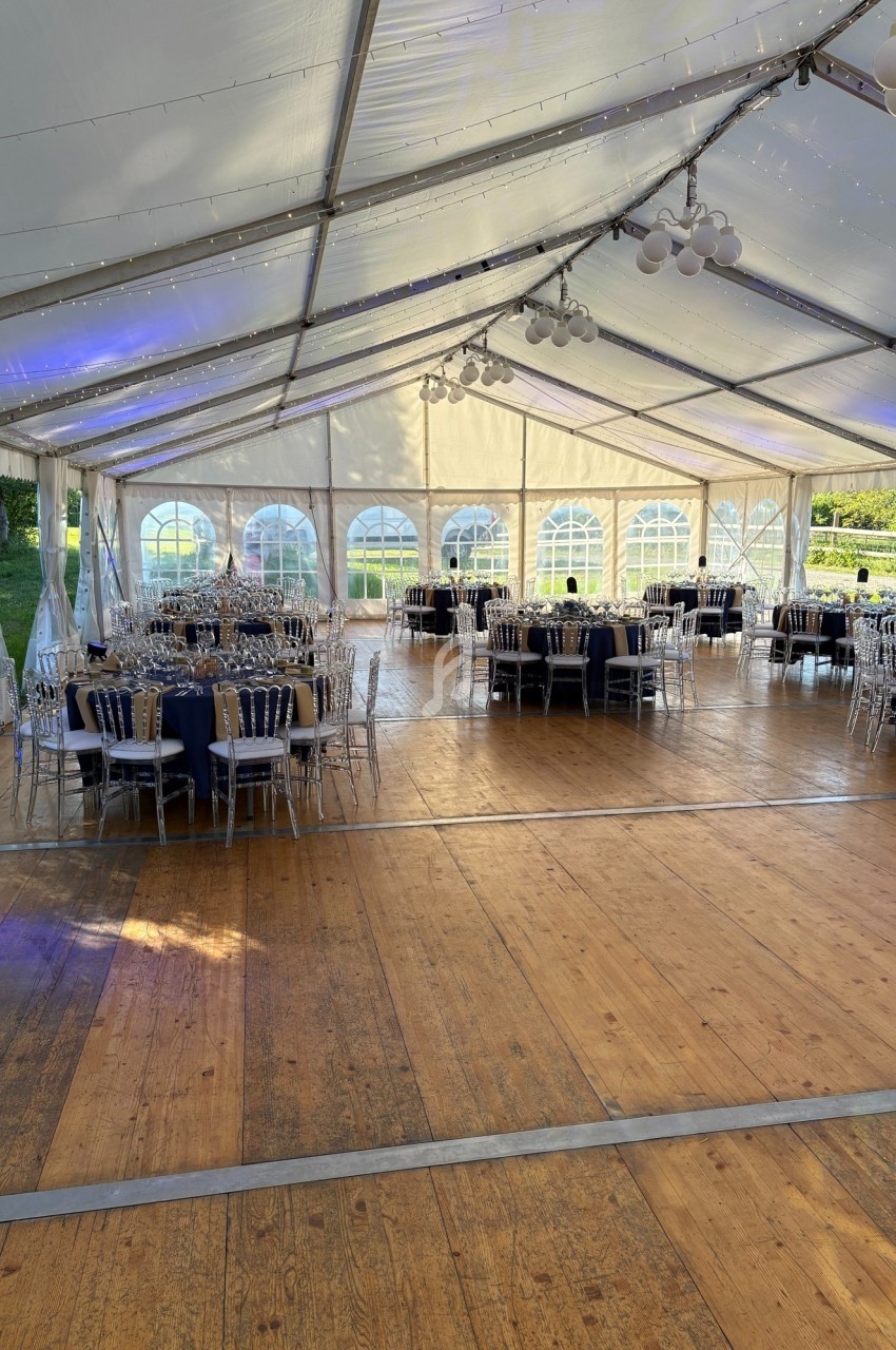 Intérieur d'une tente événementielle avec tables rondes dressées, chaises transparentes et sol en bois.