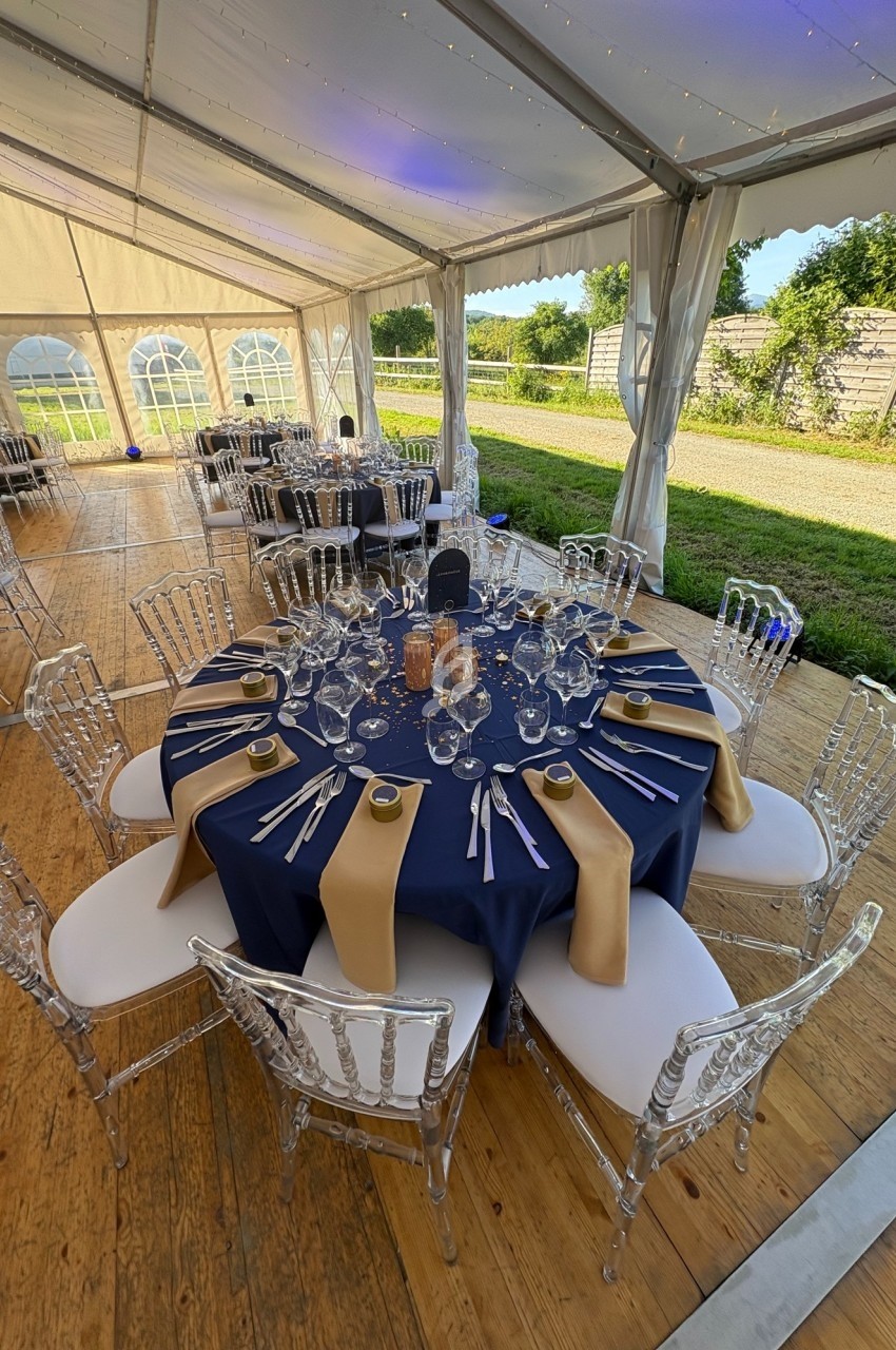 Table dressée avec une nappe bleue, serviettes dorées, vaisselle élégante et chaises transparentes sous une tente lumineuse.