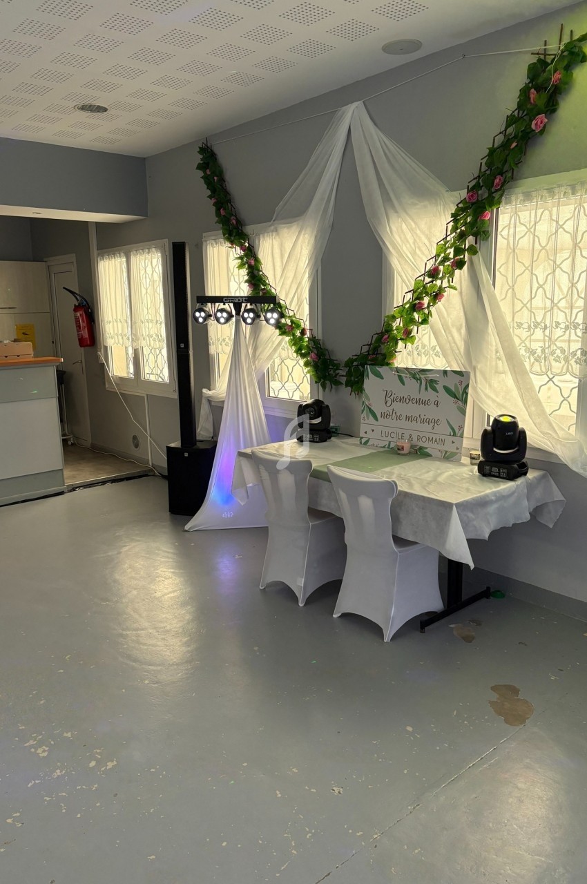 Salle décorée pour un événement avec nappes blanches, guirlandes florales et équipements d'éclairage sur des tables.