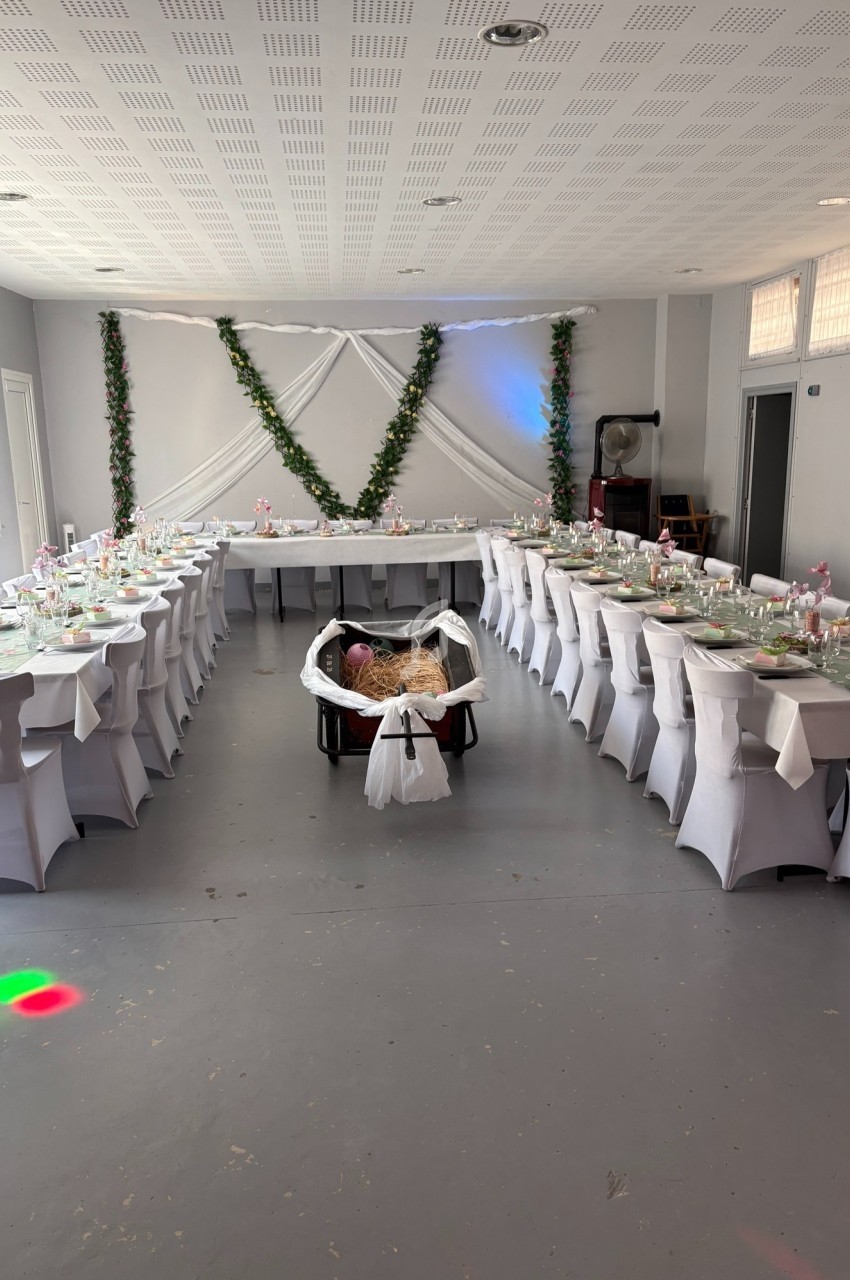 Salle décorée pour un événement, avec tables alignées, nappes blanches et guirlandes végétales sur le mur.
