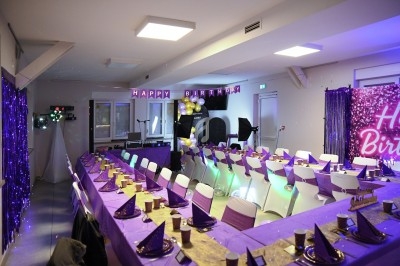 Salle décorée pour un anniversaire avec tables en U, nappes violettes, chapeaux pointus et guirlande ’Happy Birthday’.