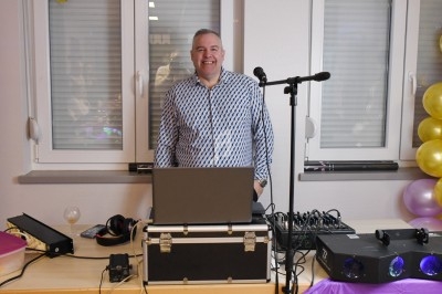 Homme souriant debout derrière une table avec équipement de DJ, micro et ordinateur, dans une salle décorée.