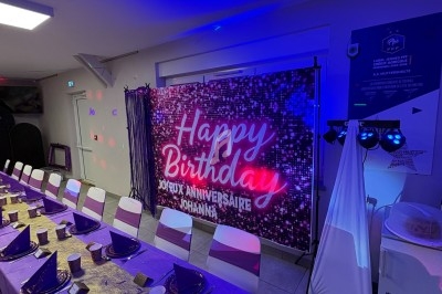 Salle décorée pour un anniversaire avec une longue table dressée en violet et un panneau lumineux ’Happy Birthday’.