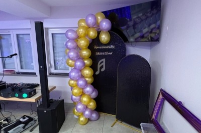 Guirlande de ballons violets et dorés décorant un coin de salle avec écran, panneaux noirs et matériel audio.