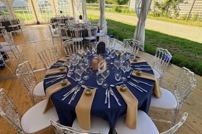 Table dressée avec une nappe bleue, serviettes dorées, verres et couverts, sous une tente lumineuse avec chaises…
