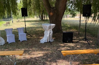 Espace extérieur aménagé pour une cérémonie avec bancs en bois, pupitre décoré et haut-parleurs sous un arbre.