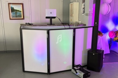 Pupitre de DJ avec éclairage coloré, haut-parleur vertical et matériel audio dans une salle sobrement décorée.