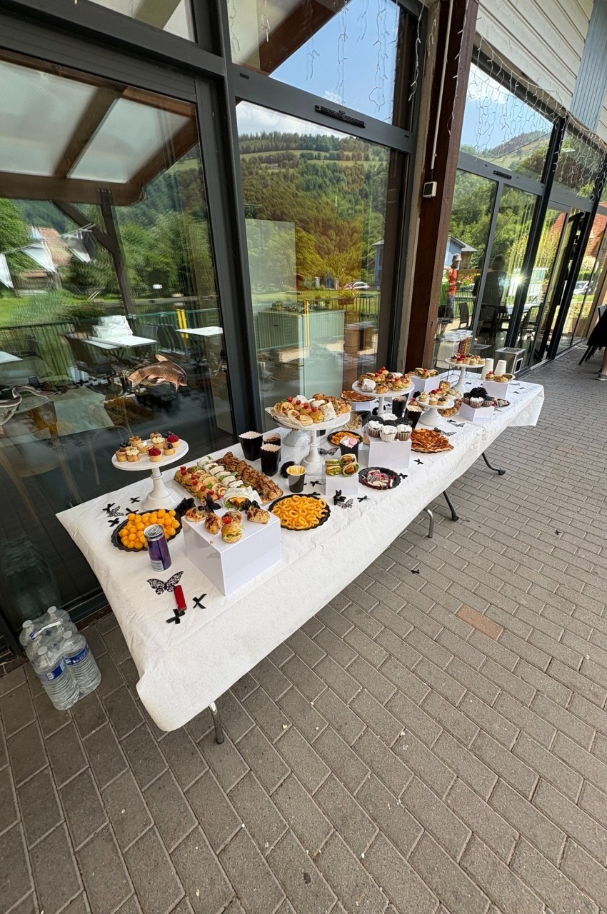 Table garnie de desserts variés et apéritifs, disposée en extérieur devant une baie vitrée avec vue sur la nature.