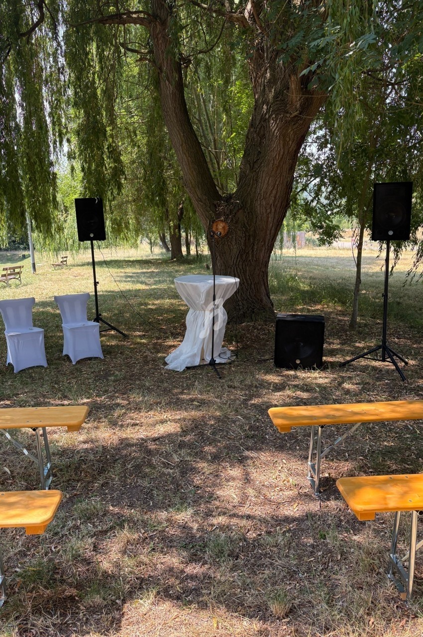 Espace extérieur aménagé pour une cérémonie avec bancs en bois, pupitre décoré et haut-parleurs sous un arbre.