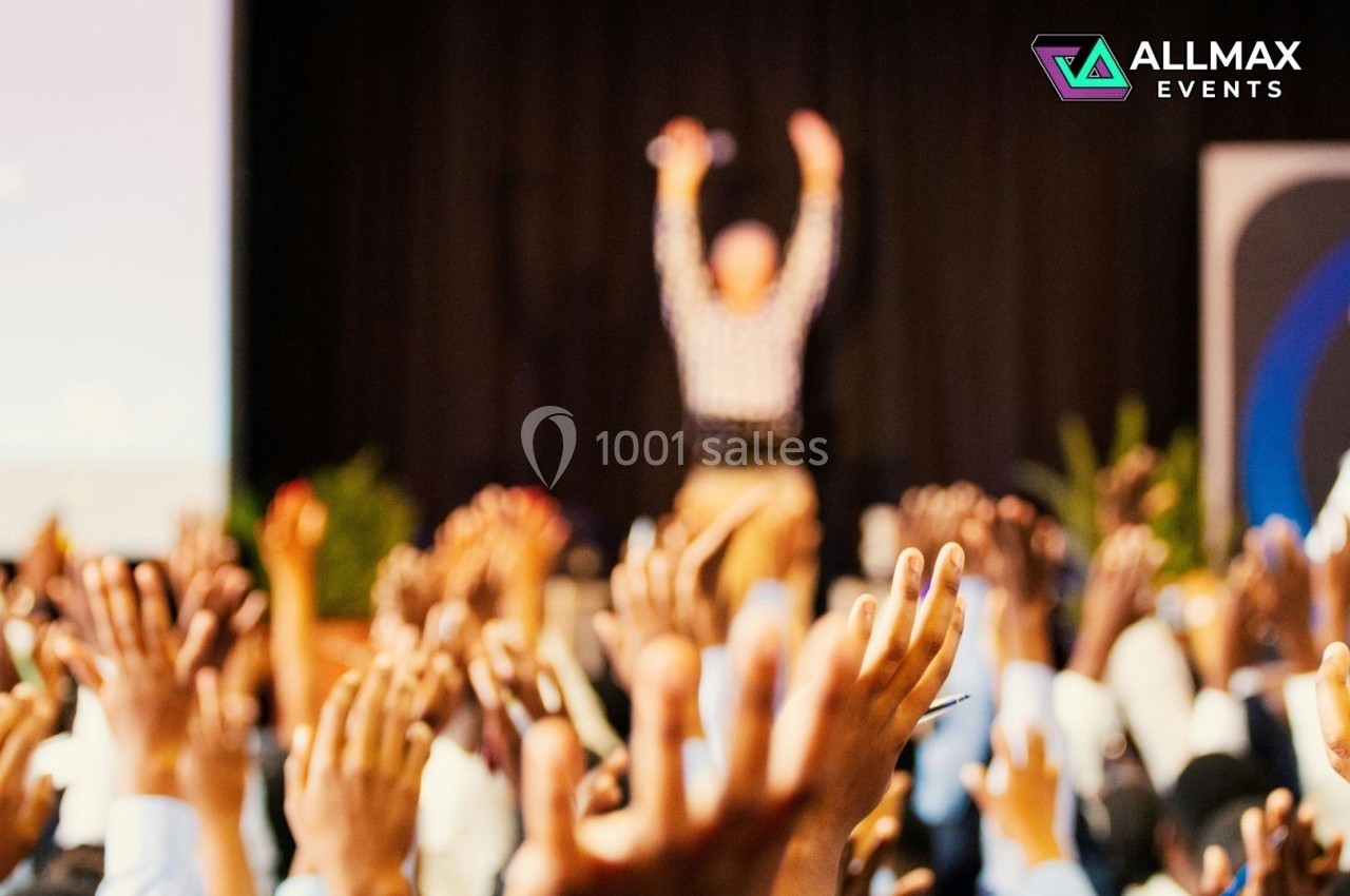 Salle comble avec des participants levant les mains, un intervenant flou en arrière-plan sur une scène.