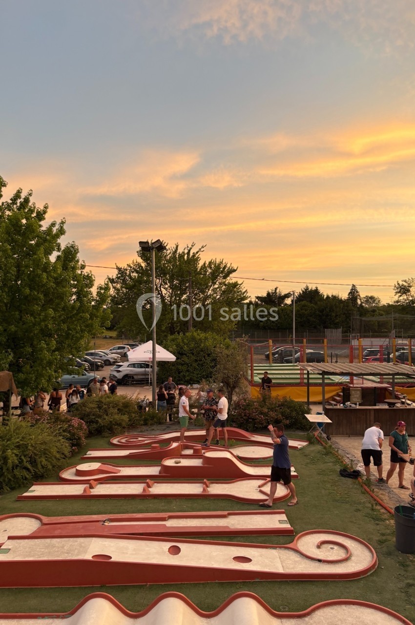 Personnes jouant au mini-golf en plein air au coucher du soleil, avec des arbres et un parking en arrière-plan.