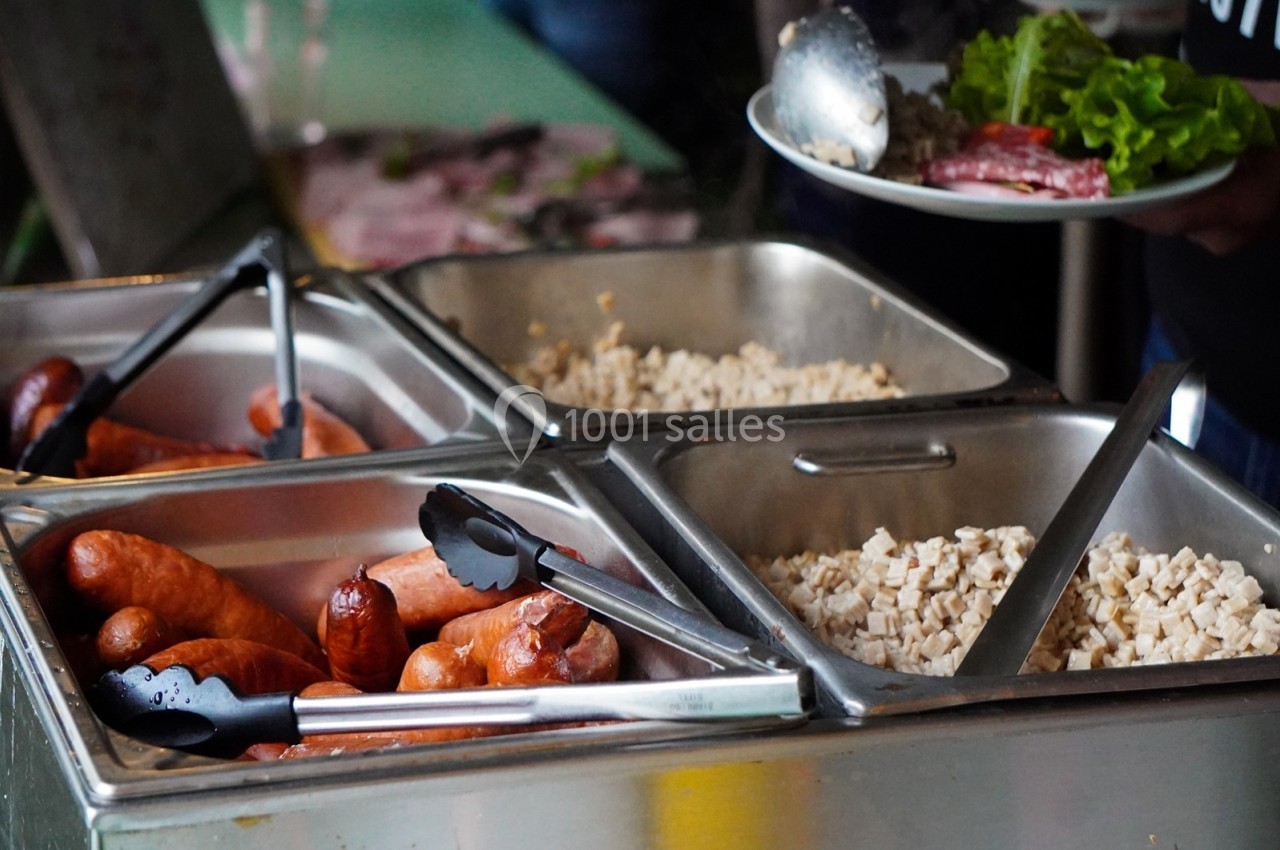 Buffet avec bacs en inox contenant saucisses, cubes de fromage et légumes, une personne se sert une assiette.