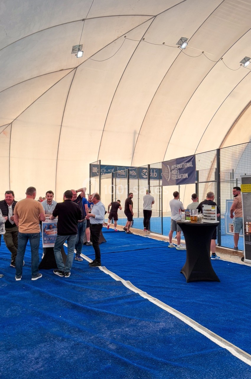 Des personnes discutent et jouent au padel dans un espace couvert avec un sol bleu et des stands promotionnels.