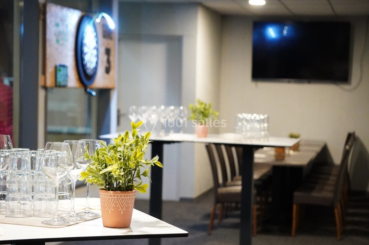 Salle aménagée avec des tables hautes, des plantes en pot, des verres et un jeu de fléchettes au mur.