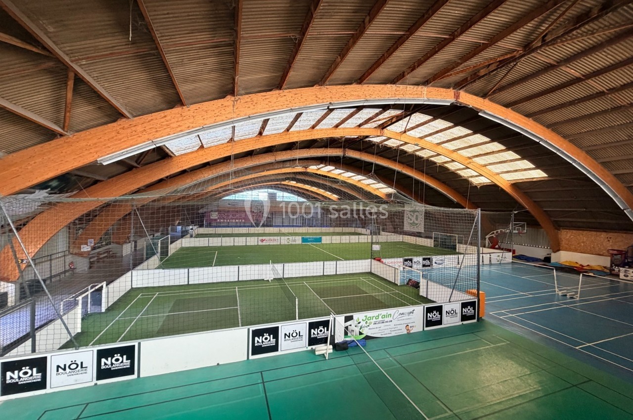 Vue intérieure d'un complexe sportif avec terrains de football en salle et de badminton sous une structure en bois.
