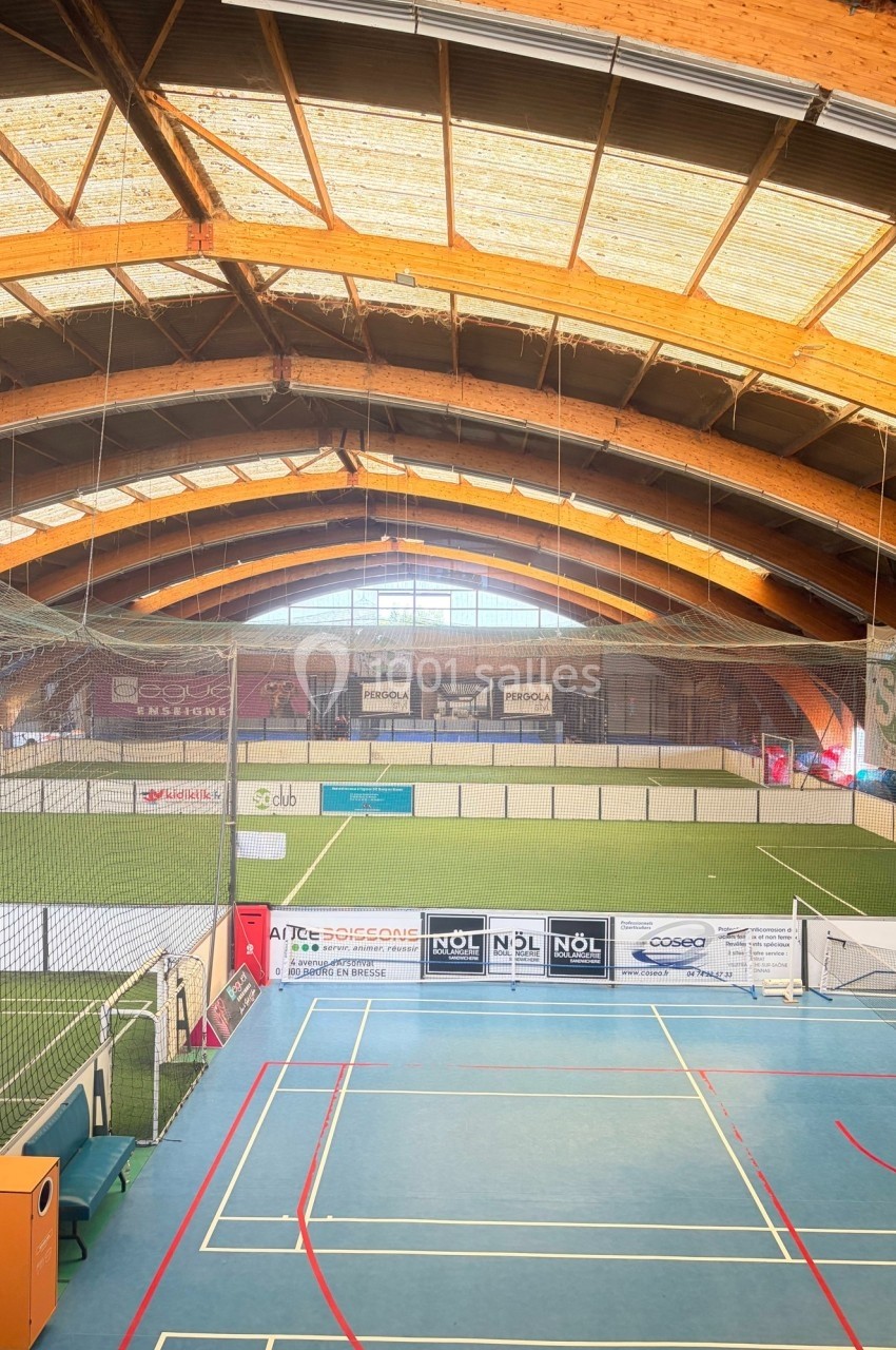 Vue intérieure d'un complexe sportif avec terrains de badminton au premier plan et terrains de football en arrière-plan.