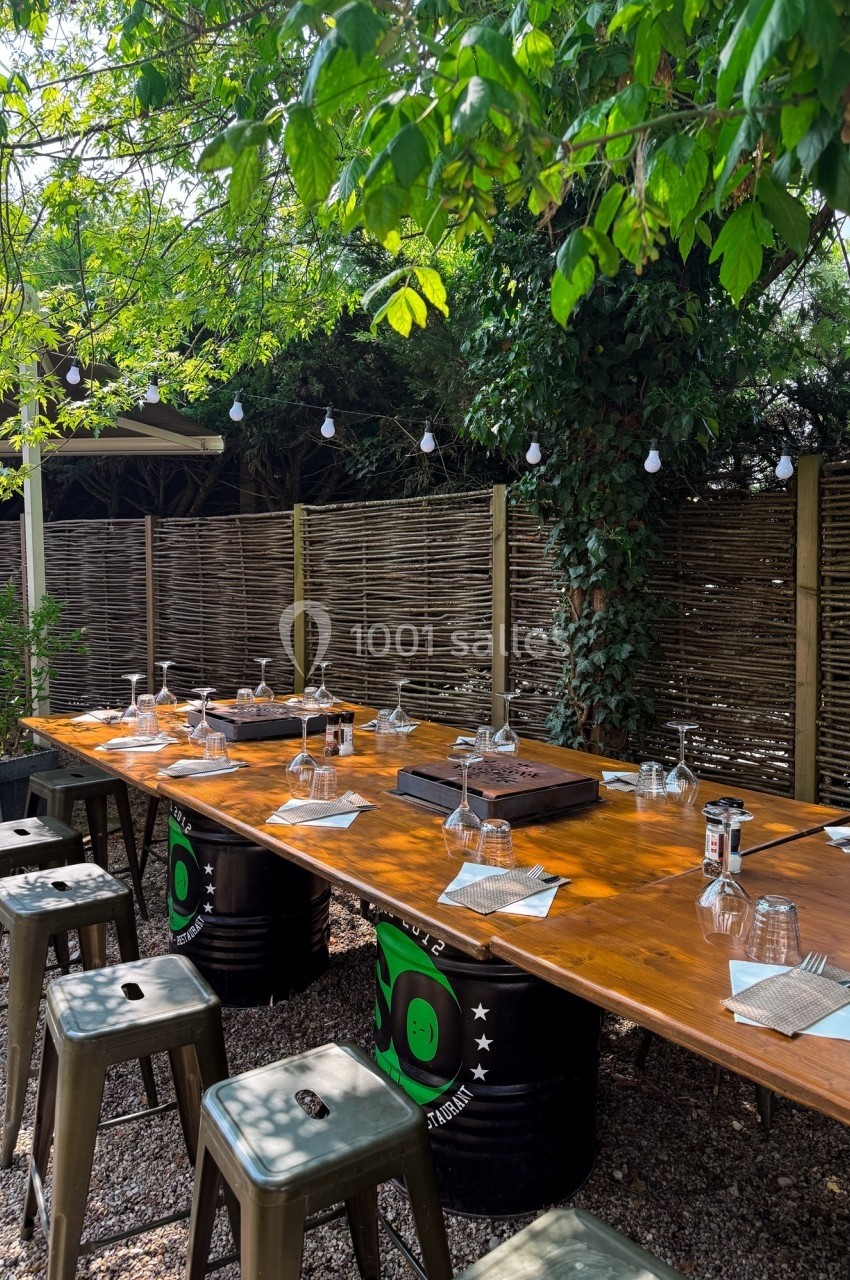 Table en bois dressée avec des verres et serviettes, entourée de tabourets, dans un jardin ombragé avec guirlandes…