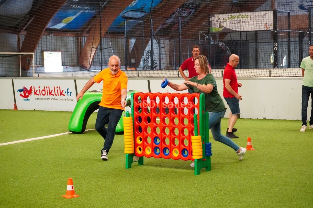 Deux personnes jouent à un jeu de puissance 4 géant sur un terrain intérieur, observées par d'autres adultes.