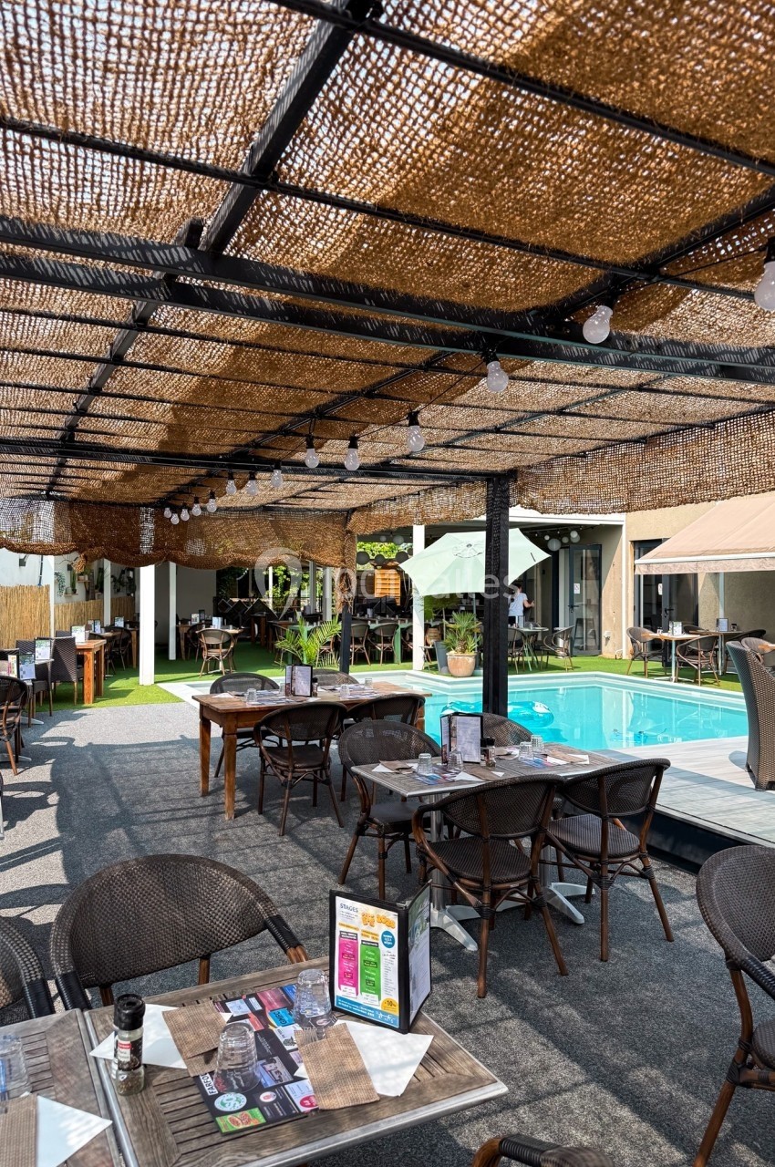 Terrasse d'un restaurant avec tables dressées, pergola en toile tressée et piscine adjacente sous un ciel dégagé.