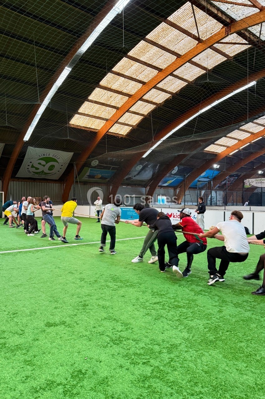 Des personnes participent à un jeu de tir à la corde sur un terrain en gazon synthétique dans un espace couvert.
