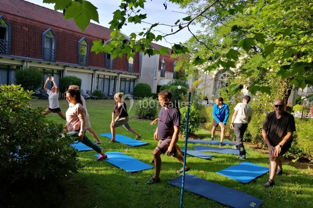 Personnes pratiquant des exercices de yoga en plein air sur des tapis, dans un jardin entouré de verdure et de bâtiments.